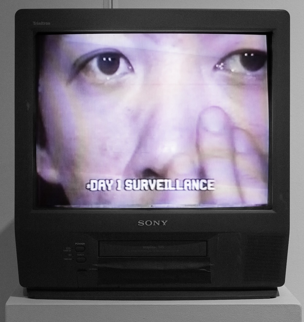 A small black Sony television set depicts a close-up black and white shot of Paul Wong’s face reaching a hand up his left cheek. The caption at the bottom of the screen reads 'Day 1 Surveillance.'