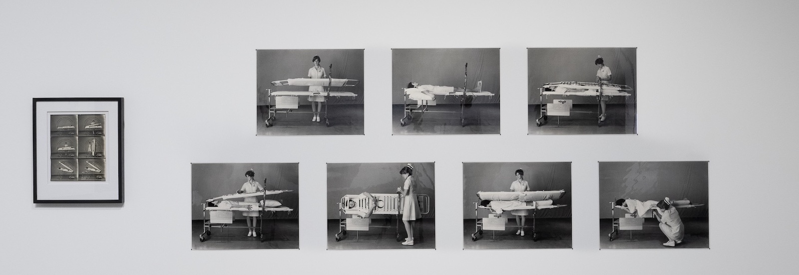 A collection of black and white photographs are mounted on a white art gallery wall. They depict a person in a white nurse’s uniform working with a patient (the artist Theodore Sasketche Wan) in a stretcher-like contraption.