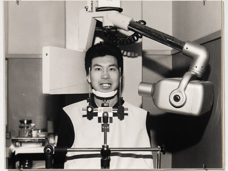 A black and white photograph of Theodore Sasketche Wan, a Chinese Canadian man with short black hair facing the camera, seated in a chair and receiving a dental X-ray.