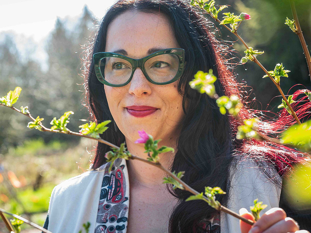 Jennifer Grenz is an Indigenous woman with long dark hair, light skin and green glasses. She stands behind a blossoming stand of salmonberry branches with green leaves and small pink flowers. The sun is shining.