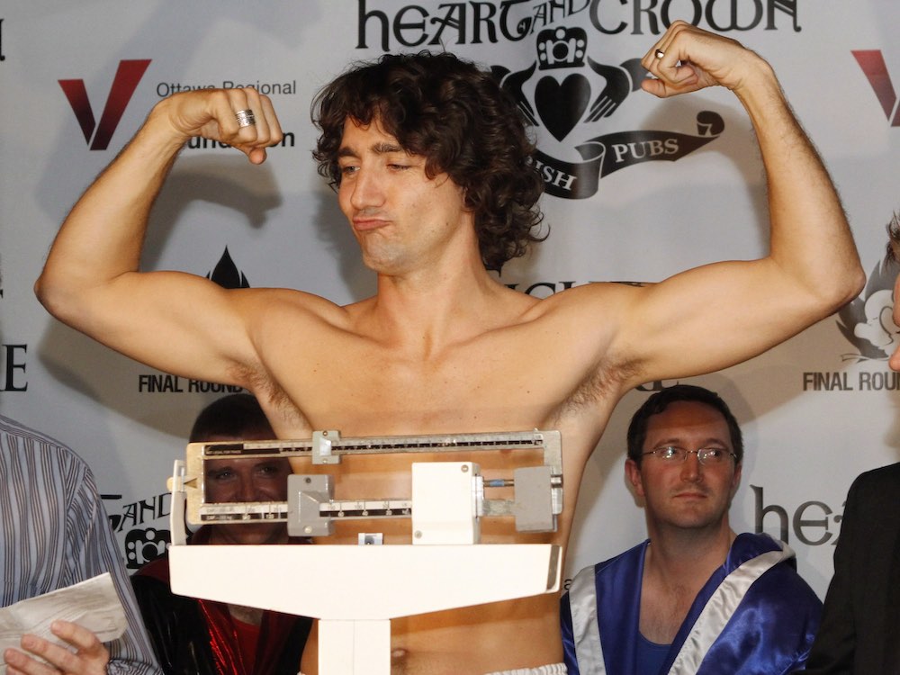 Justin Trudeau has light skin and curly brown hair. He is wearing white boxing shorts without a shirt, and strikes a muscle-man pose while standing on a white scale during a boxing weigh-in. Behind him is a man in a blue boxing robe.