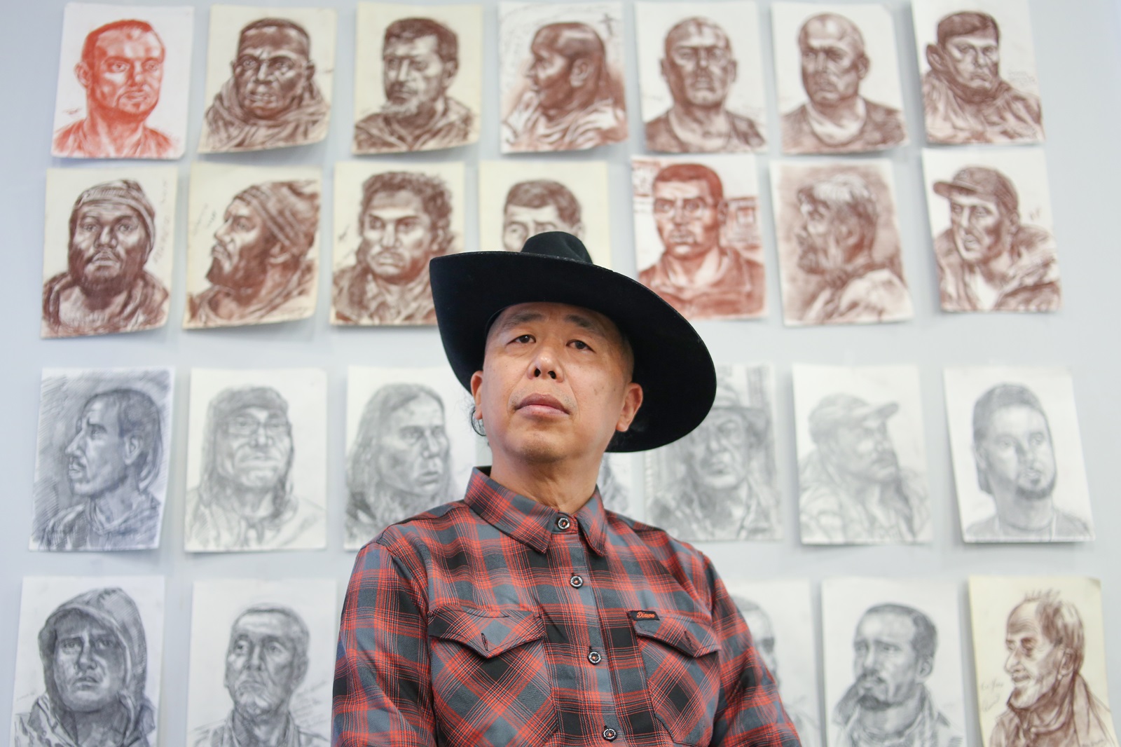 A Chinese man in a black cowboy hat and red plaid shirt is seated against a white wall where rows of charcoal portraits are placed. The sketches are in red, brown and grey.