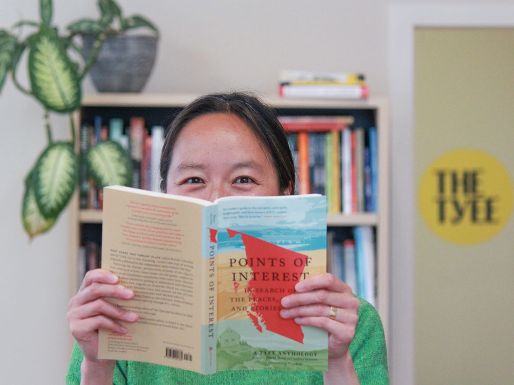 Tyee senior editor Jackie Wong poses in The Tyee office, with the book 'Points of Interest' half covering her face. She is smiling from behind the book.