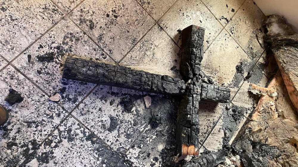 A charred wooden cross lies on a tile floor surrounded in black ash.