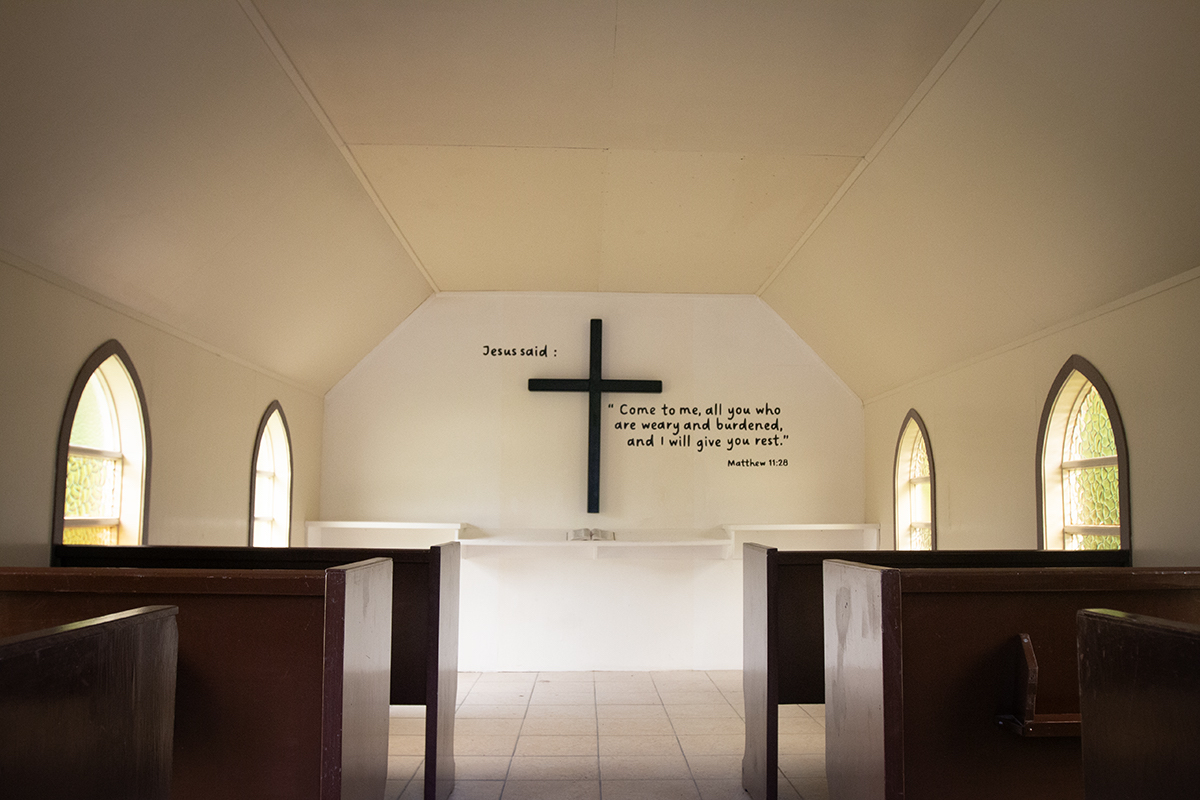 The white interior of a chapel with sloped ceiling and dark wooden benches on either side. On the back wall is a cross and the message 'Jesus said: Come to me, all you who are weary and burdened, and I will give you rest. Matthew 11:28.'