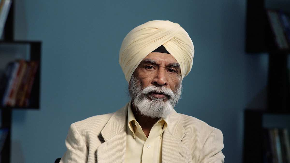 An older man with medium skin and a grey beard wears a cream-coloured corduroy blazer and matching cream-coloured turban. He is seated against a blue background and wears a serious expression mid-speech.