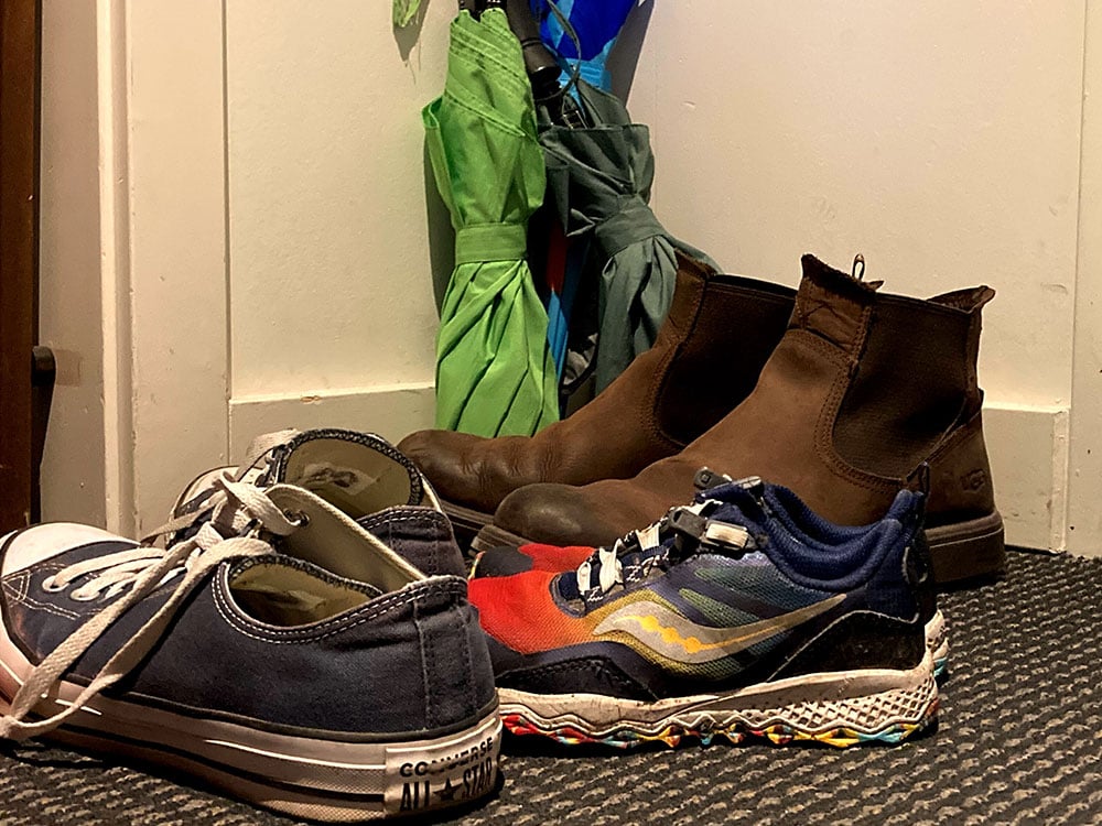 From left to right, a pair of navy Converse All Stars, a colourful pair of children’s running shoes and a brown pair of Chelsea boots stand in the corner of an interior space with three umbrellas behind them. To the right is an apartment door painted blue.