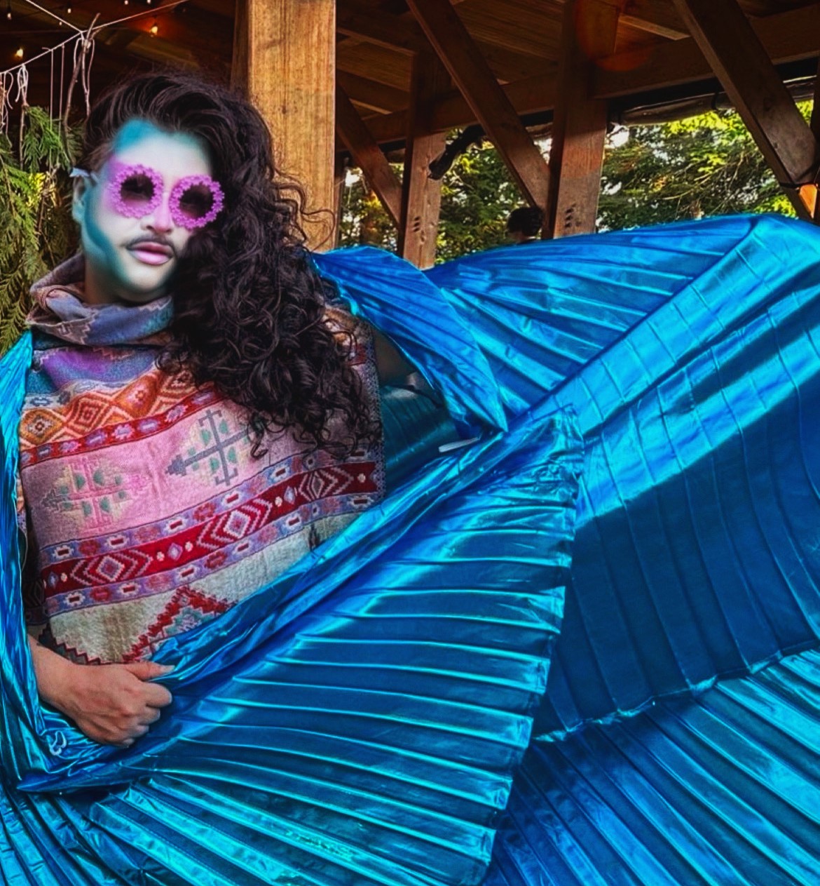 A person with medium-light skin tone, long, curly dark brown hair, blue and white face paint, and pink flower sunglasses, faces the camera and holds out a long shiny blue shawl.