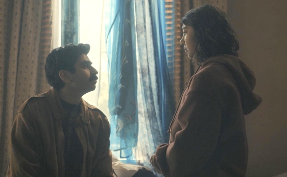 A young man with short wavy dark hair, a moustache and medium skin looks up at a young teen girl to his right, standing in a brown hoodie with short wavy dark hair and medium skin. They are indoors near a curtained window.