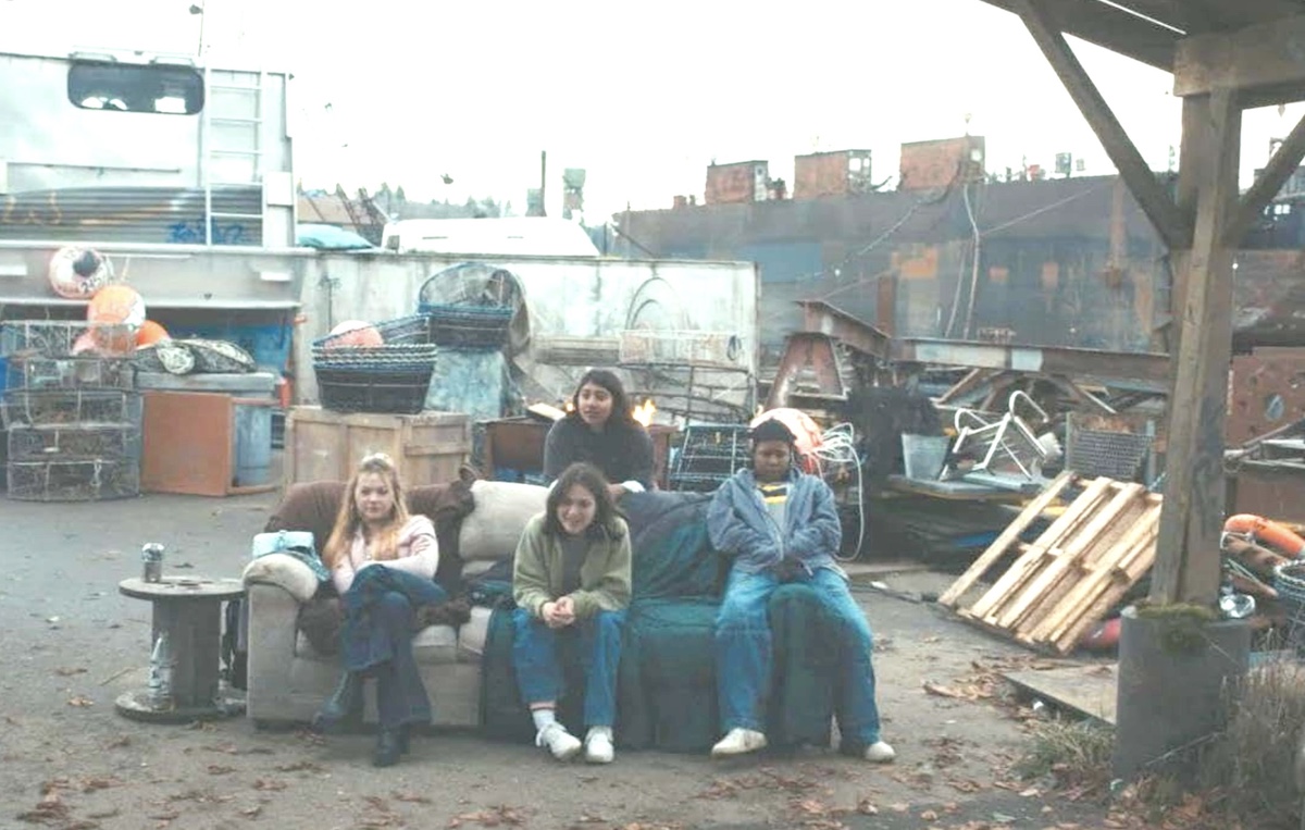 A group of four actors in their early teens sit on a worn grey sofa covered in blankets in a junkyard outdoors. Three girls sit across the couch and one girl with medium skin and dark hair hangs over the back. Around them are piles of discarded items like furniture and wood pallets. The sky is white.