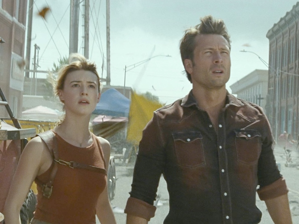 A film still depicts a woman, left, in a red tank top with light skin and blond hair tied back. To her right is a man with light skin, short blond hair and a dark button-down shirt. They are standing outside amidst stormy rubble that is blowing about. They are looking concerned as they gaze into the middle distance.