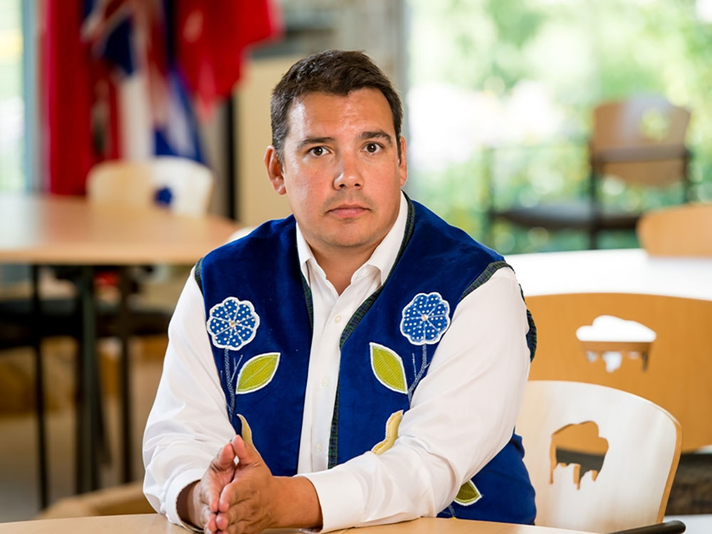 Niigaan Sinclair is seated at a table. He has short dark hair, medium skin and is wearing a blue floral vest over a white button-down shirt. His hands are pressed together and he is looking directly at the camera.