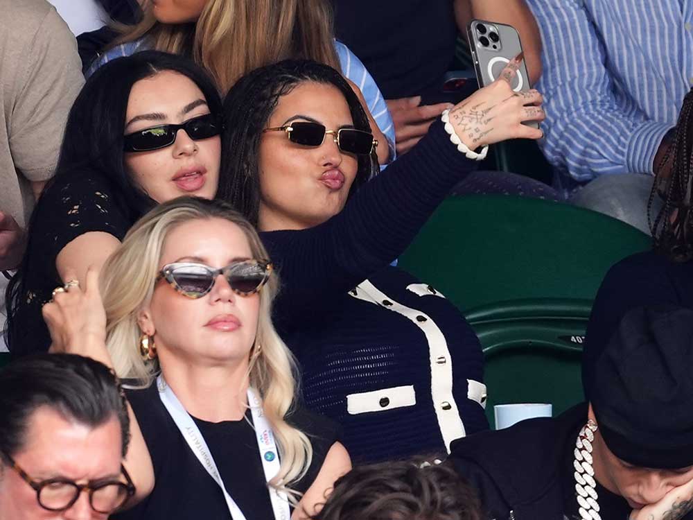Two young women with long dark hair and sunglasses poses for a selfie while seated in a crowded set of bleachers at the Wimbledon Championships.