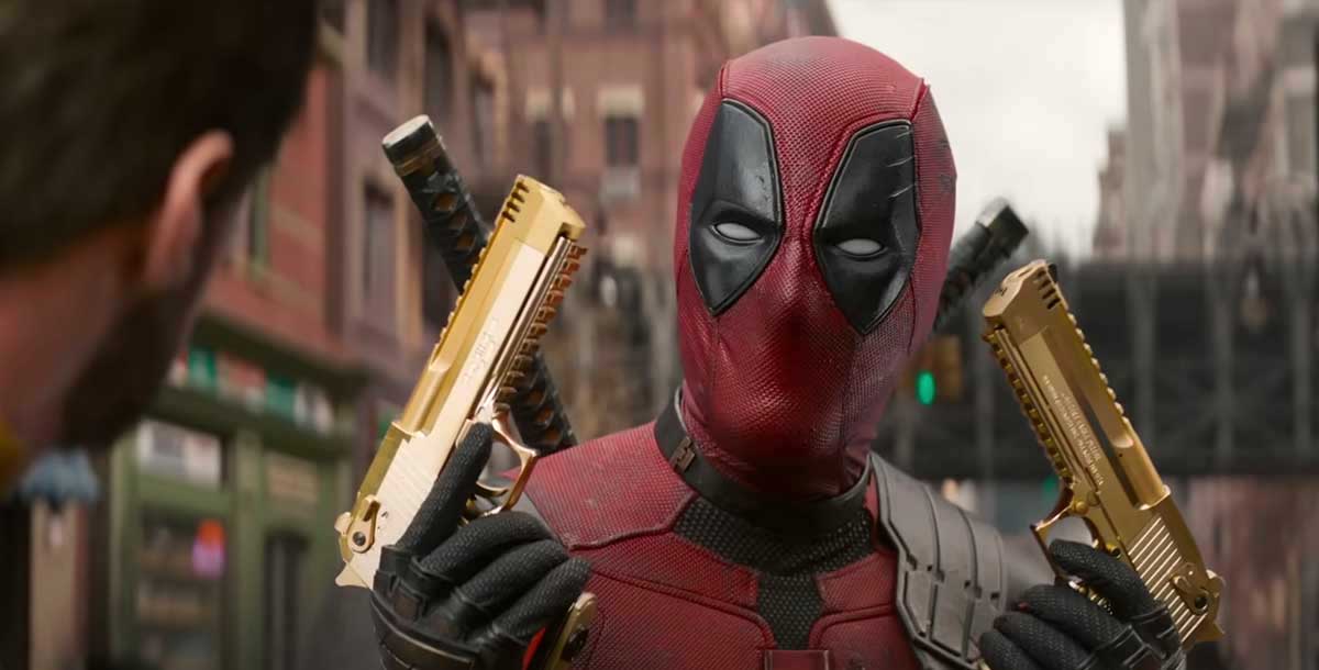 An actor in a red and black superhero bodysuit that covers his face stands outside in a downtown street. He wields two gold pistols, pointing each of them up using both hands.