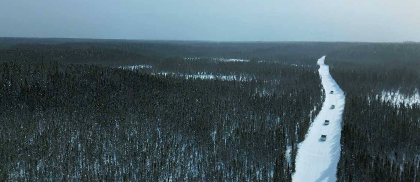 Drone footage captures a small line of vehicles on a remote snowy road flanked by forest.