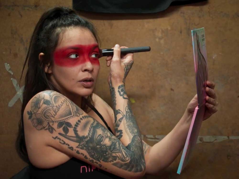 Sage Morin is an Indigenous woman with her long dark hair tied back as she applies red paint in a band across her eyes. She holds a hand mirror to guide her progress while seated against a worn plywood wall.
