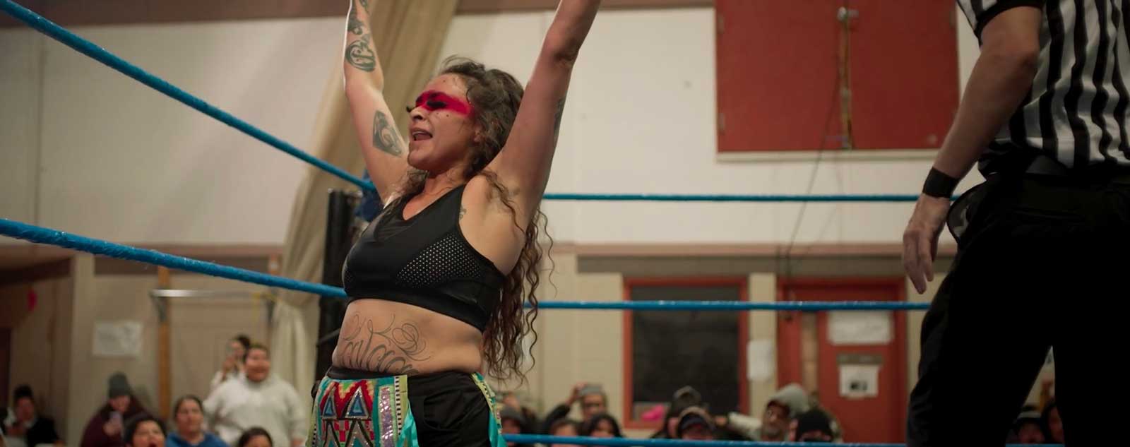 Sage Morin is an Indigenous woman with long dark wavy hair. She has painted a band of red across her eyes. She wears a black sports bra and wrestling shorts with Indigenous designs on the front. She stands in the middle of a wrestling ring inside a school gym with both her arms raised in victory.