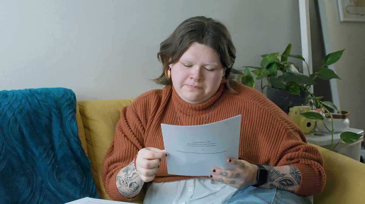 jaye simpson has short, wavy, light brown hair and light skin, and is seated on a yellow sofa in an apartment. She wears a pumpkin-coloured sweater and light blue jeans. She holds a white sheet of paper in her hand and is looking down at it, verklempt.