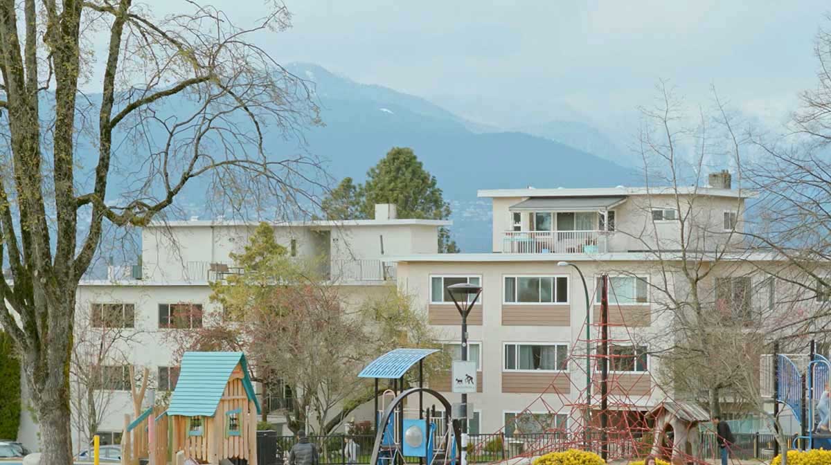 Two mid-rise apartment buildings, in white and beige, stand against a misty sky and blue mountains in the background. A playground is in front of the buildings.