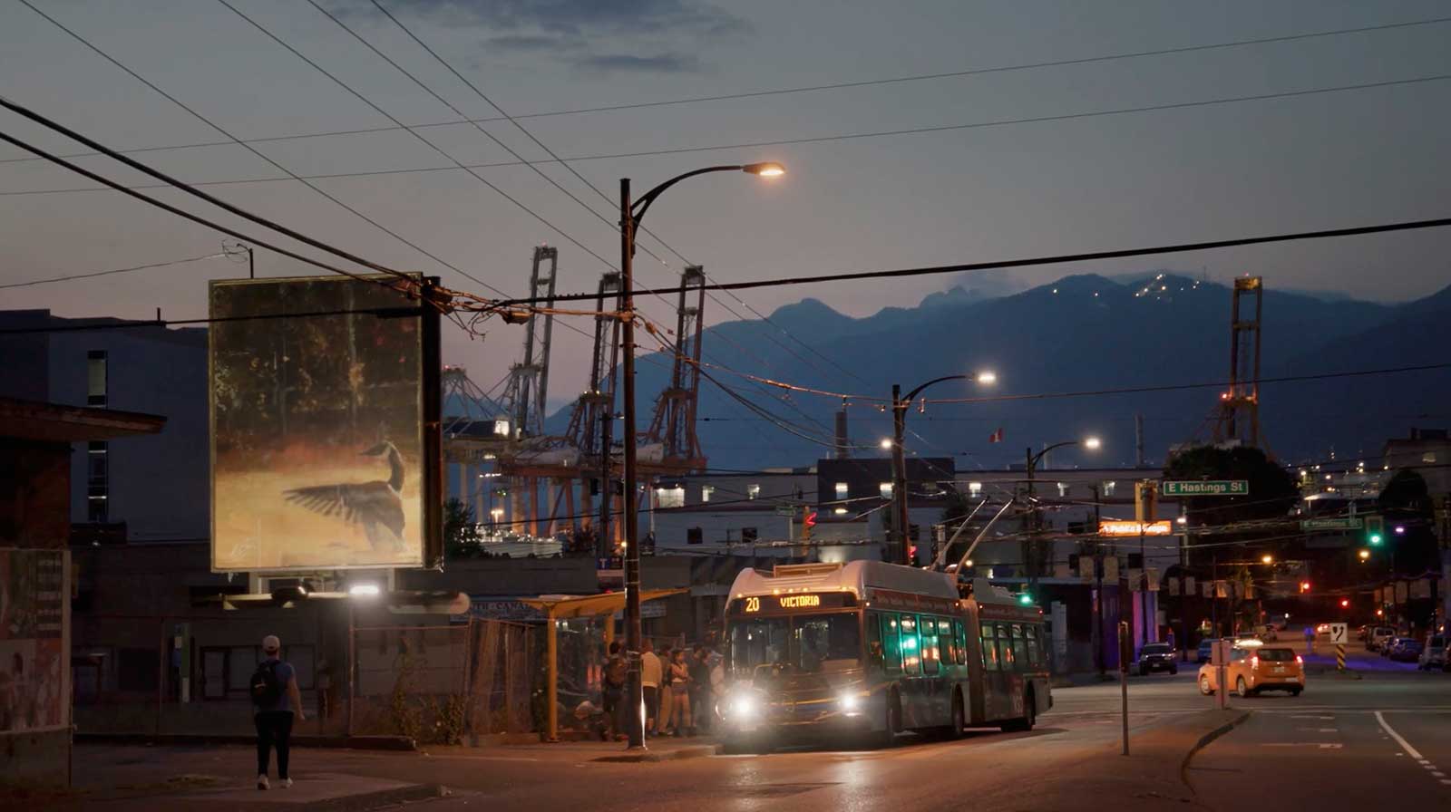 The No. 20 Victoria bus pulls up to a stop on a street in Vancouver at dusk. A billboard featuring a painting of a Canada goose is to the left of the scene. In the background are mountains, tall structures that are part of the nearby port, low buildings and street lights.