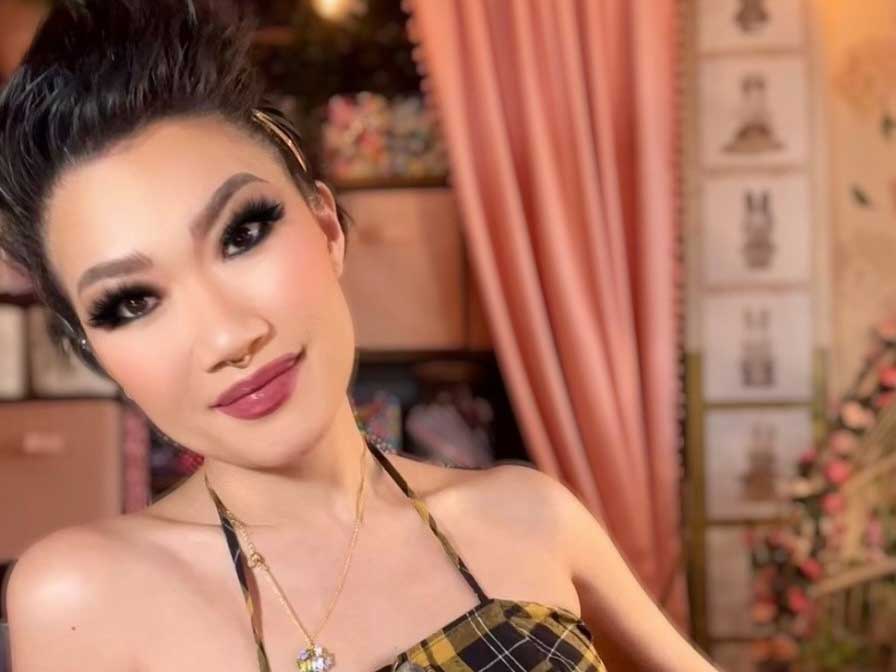 Jenny Heijun Wills is a Korean woman with her long dark hair pulled back in an updo. She wears dramatic eye makeup and has a septum piercing. She is wearing a black and yellow gingham halter dress and is seated in an interior space with a pink curtain in the background.
