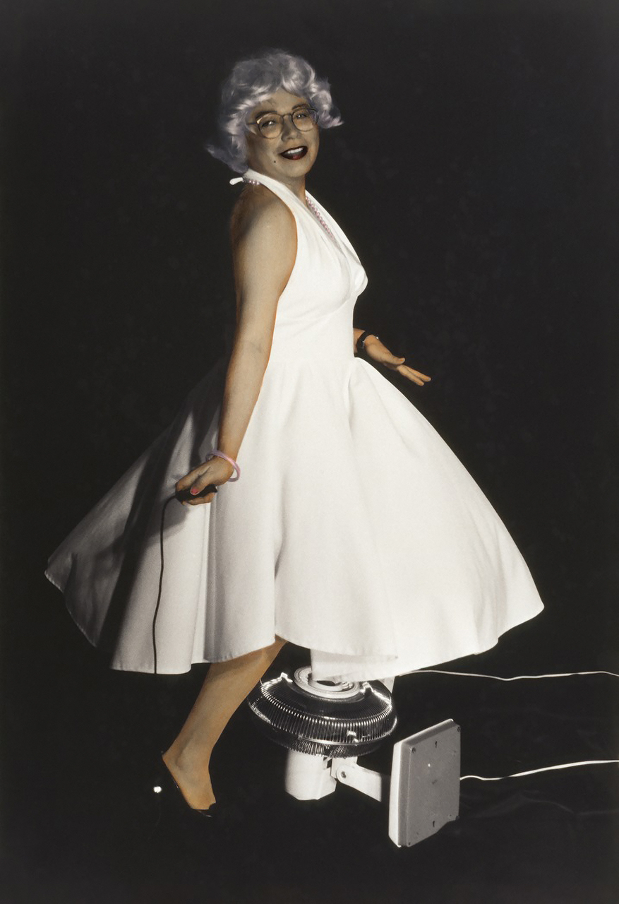 A colour photograph of an Indigenous woman in glasses with a light purple wig and white Marilyn Monroe-style dress. The woman is smiling playfully and stepping over an upturned white household fan against a black background.