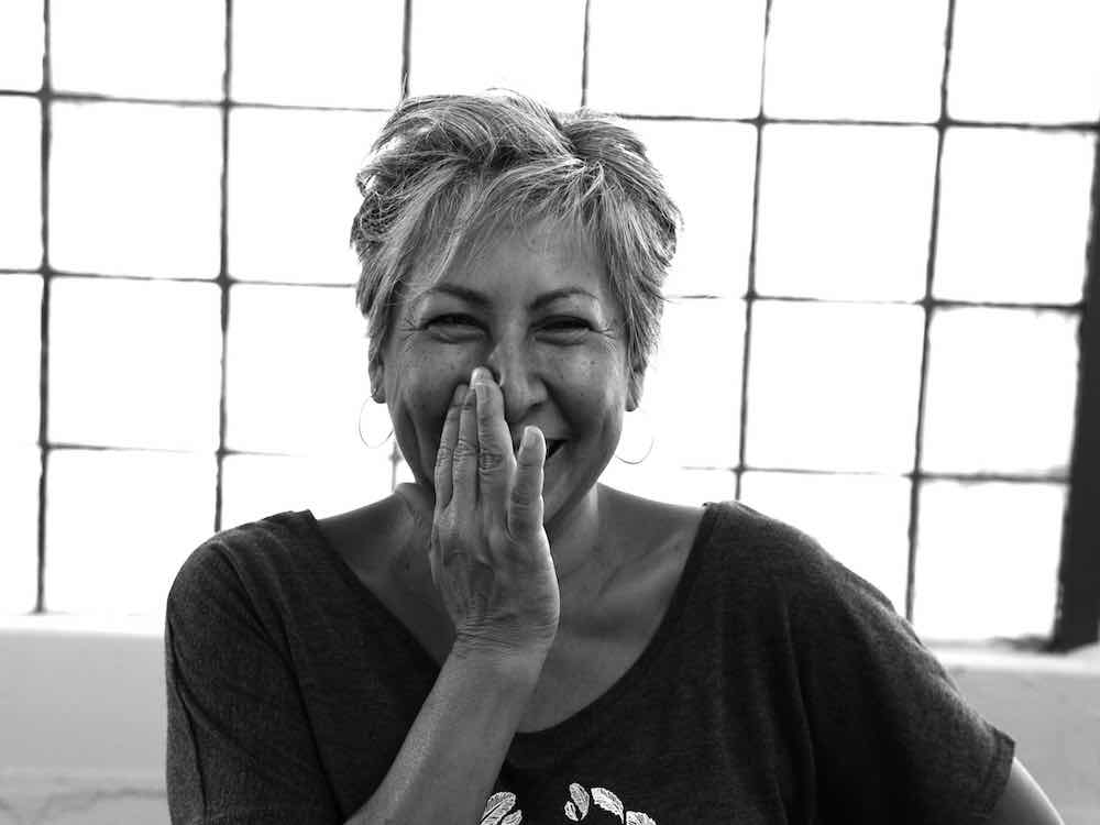 A black and white portrait of Rebecca Belmore depicts an Indigenous woman with short grey hair, mid-laugh with her hand to her face. Behind her is a window with a grid pattern.