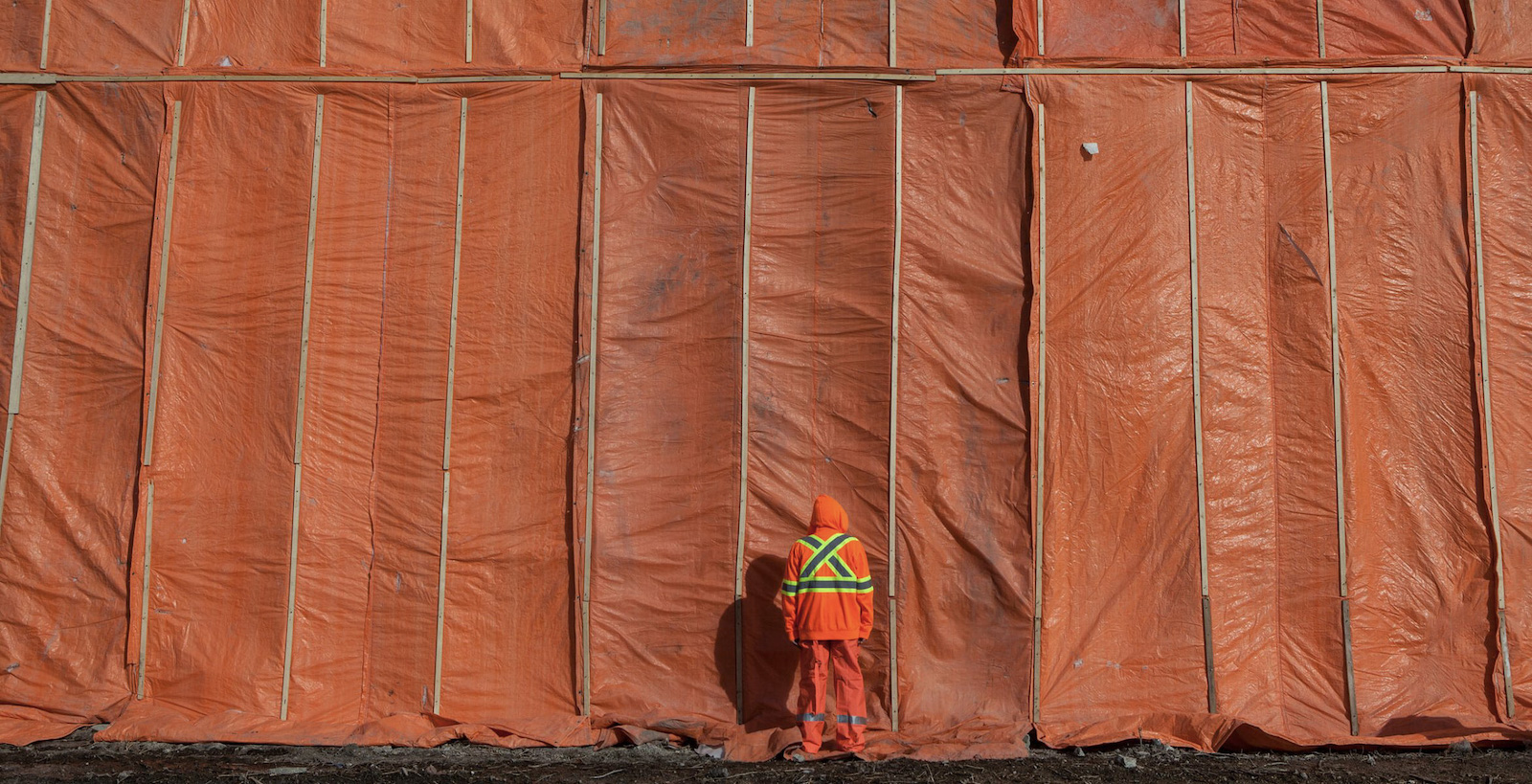 Rebecca Belmore stands with her back turned to the camera. She is wearing bright orange safety gear with a fluorescent yellow 'X' on her back. She stands against a wall of orange tarpaulins.