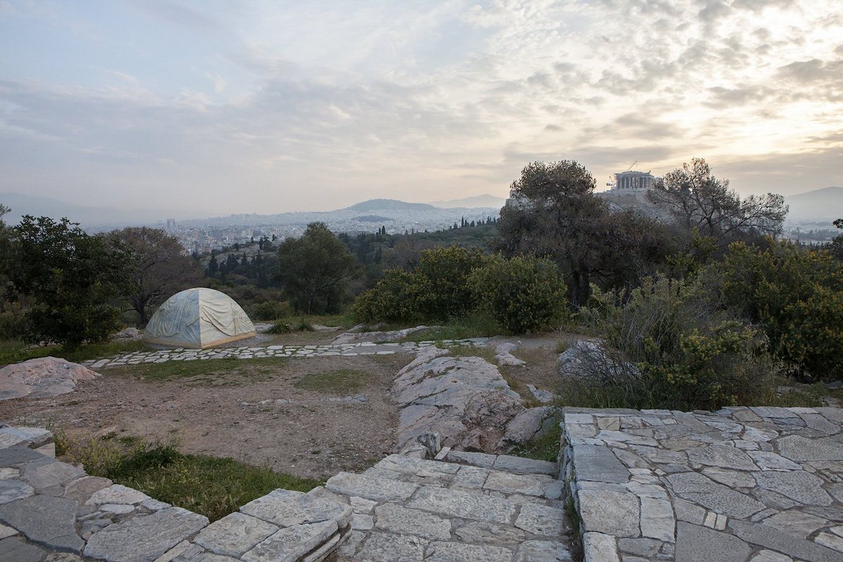 A photograph captures an expansive view of Greece with the Acropolis in the background. In the foreground is a small grey dome tent in a clearing near a walkway made of cobblestones. The sky is misty and grey.