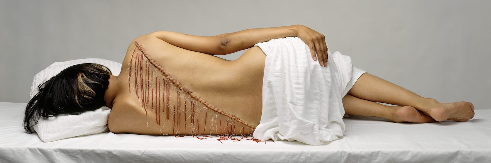 A photograph of a woman with short dark hair with her back facing the camera. She is lying on her side with a white fabric wrapped around her waist. Red weaving and string winds in a diagonal line across her bare back.