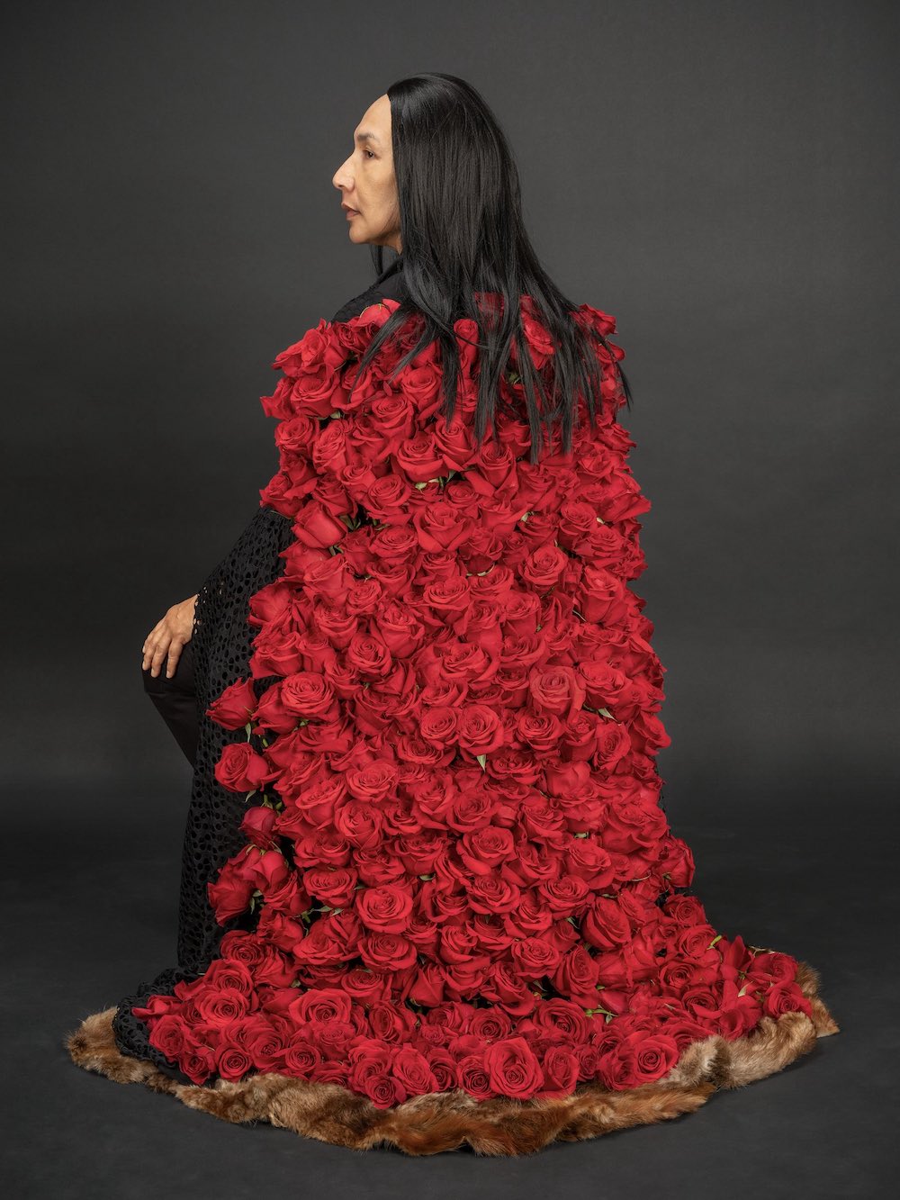An Indigenous woman with long dark hair sits with her back to the camera against a black studio background. She is wearing a floor-length shawl of red roses.
