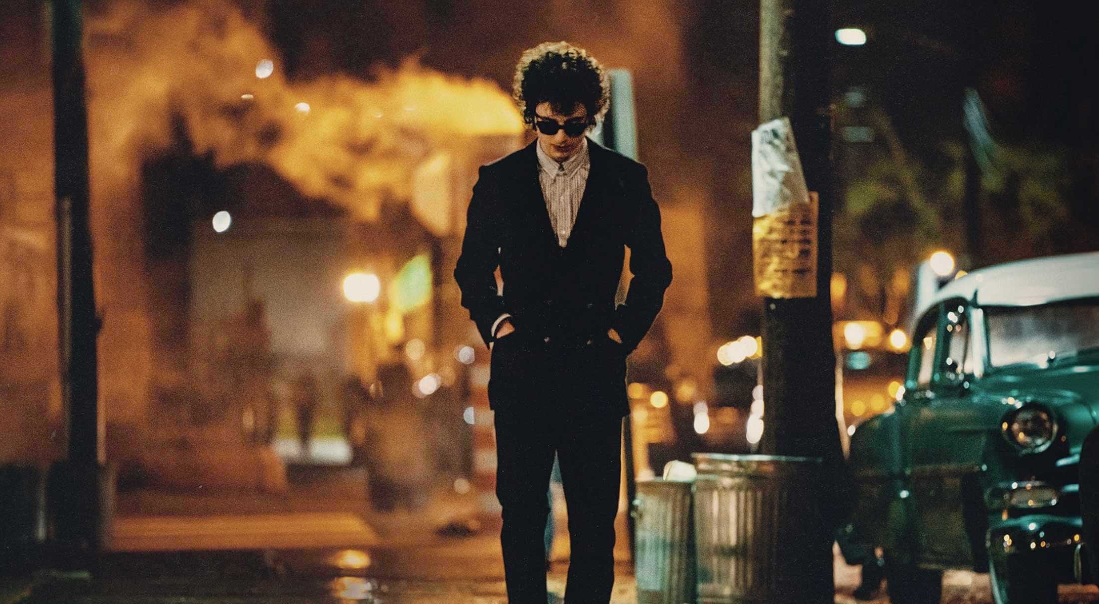 Actor Timoth&eacute;e Chalamet in 'A Complete Unknown' as Bob Dylan. He has dark curly hair and is wearing sunglasses, a dark blazer over a grey button-down shirt and dark denim jeans. He is walking down a city street at night. Grey trash cans line the street.