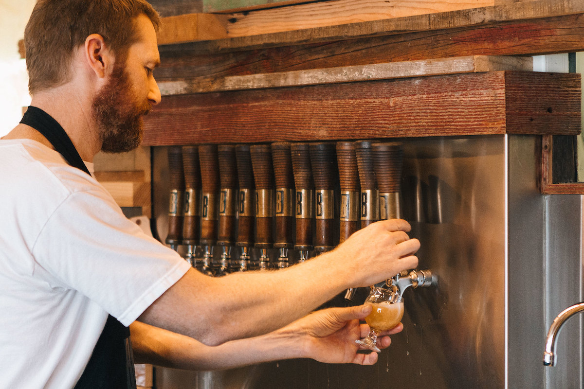 A man with reddish hair and a beard is wearing a white T-shirt under a black apron. He is standing before a row of wooden taps marked with the letter B and pouring beer into a small round glass.