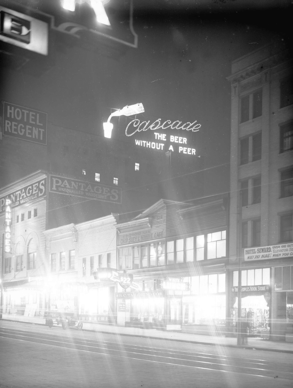 An archival black and white photo of a Vancouver streetscape at night. The white lights from the ground-floor shops of several tall buildings are refracted across the page. Above the buildings, a neon sign features a silhouette of a bottle being poured into a glass, beside a sign that reads 'Cascade: the beer without a peer.'