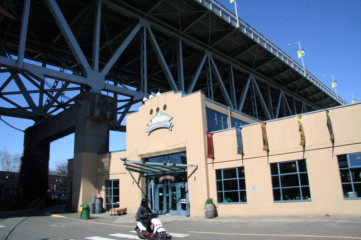 A street shot of the exterior of a light beige building under a bridge that houses Granville Island Brewing on Granville Island.