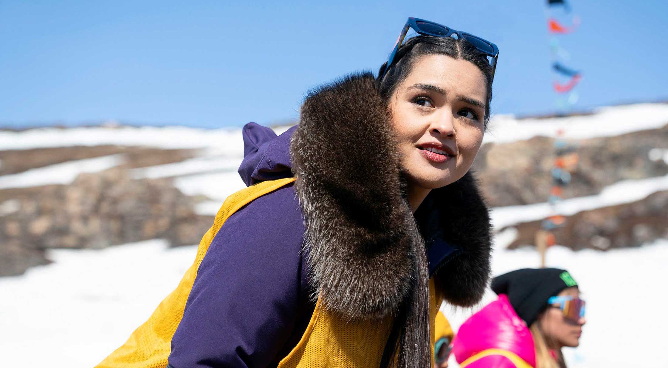 Actor Anna Lambe is outdoors in a snowy setting against a blue sky. Her long dark hair is pulled back under a yellow and purple winter layers and a large brown fur collar. A pair of black sunglasses balances atop her head. She is looking up towards the left of the frame.