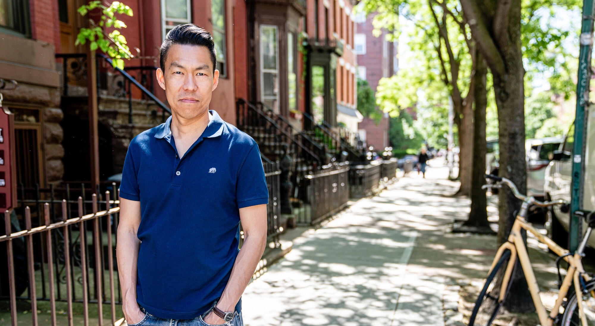 Jack Wang has short black hair and is standing on a street lined with brownstone apartment buildings on a sunny day. He is wearing a blue collared short-sleeved shirt with his hands in the pockets of his blue jeans. A yellow bike leans against a green pole to the right of the frame.