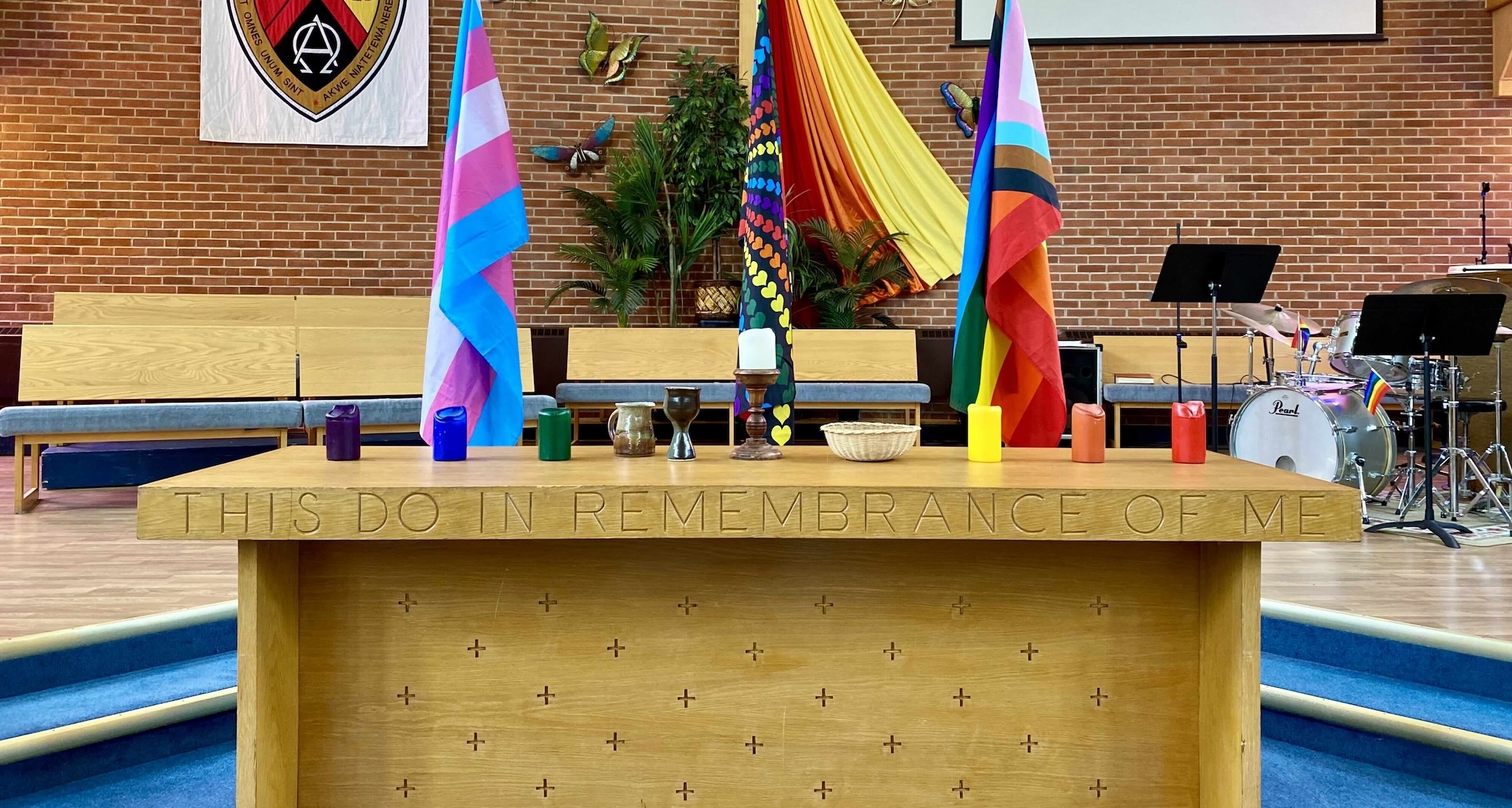 The interior of Knox United Church features a long wooden table with the inscription 'This do in remembrance of me' inscribed on the side of its ledge. Behind the table are colourful flags for trans and queer pride. The table is topped with rainbow-coloured candles. In the background is a wooden cross against a brown brick wall and a drum kit.