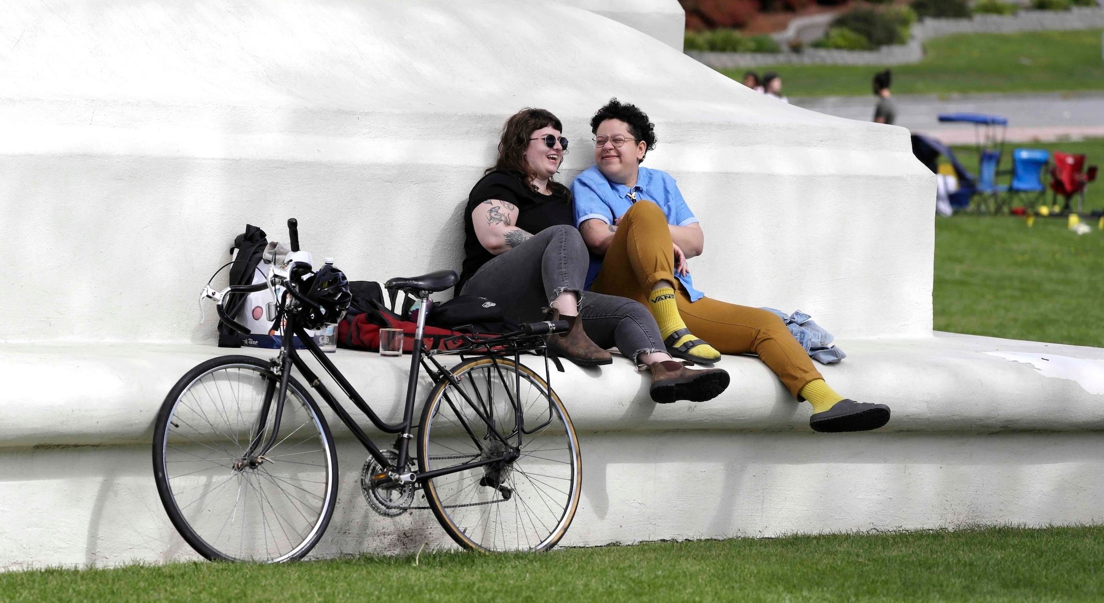 A bicycle leans against the base of a large white marble monument upon which two people sit together on its lower ledge. Hannah Smith, left, has shoulder-length dark hair, pale skin, sunglasses and a black T-shirt with dark grey jeans. Jabree Robinson, right, has short dark curly hair, medium-light skin, a short-sleeved blue shirt and tan trousers. It’s a spring day and people are gathering with lawn chairs in the background.