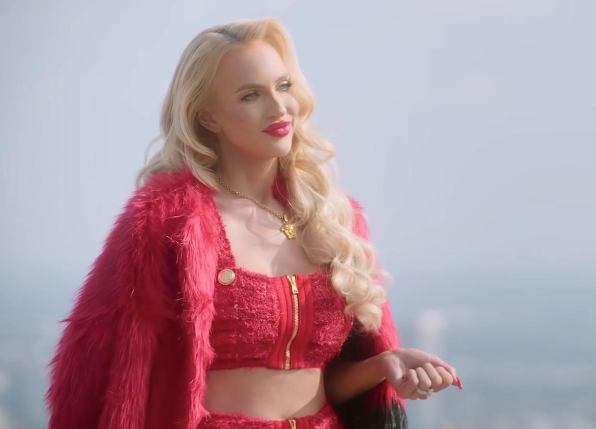 Christine Quinn has long wavy blond hair and pale skin and is wearing a glamorous red outfit adorned with fur and gold accessories. She is standing outdoors against a hazy sky.