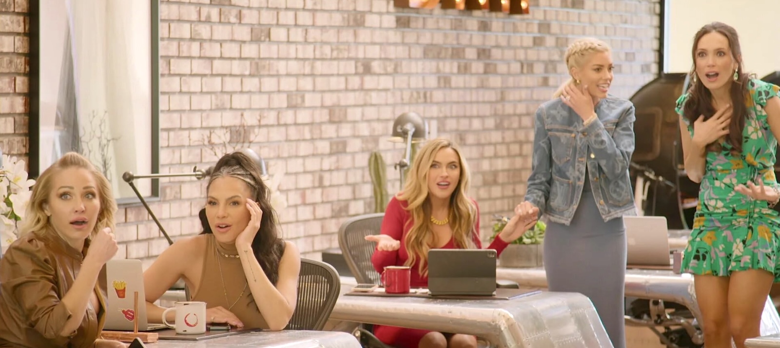 A group of women sit at desks or are standing in a luxurious office environment with brick walls, each of them looking off camera with incredulous expressions.