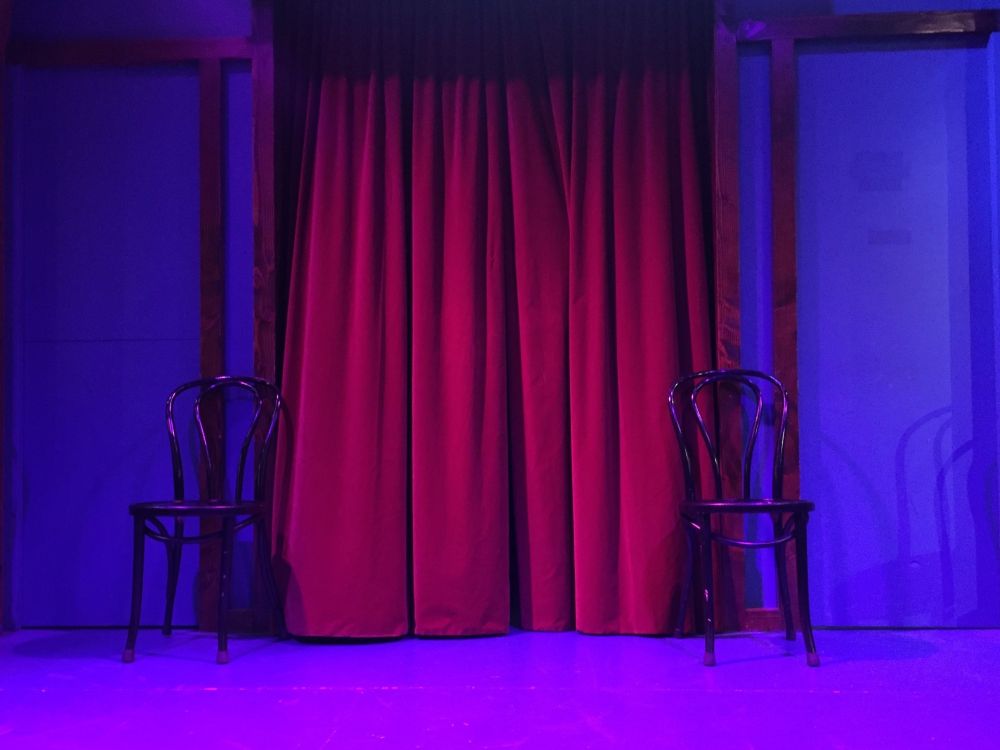 A small theatre stage features red curtains and two wooden chairs, each placed on opposite sides of the stage. The stage is cast in blue light.