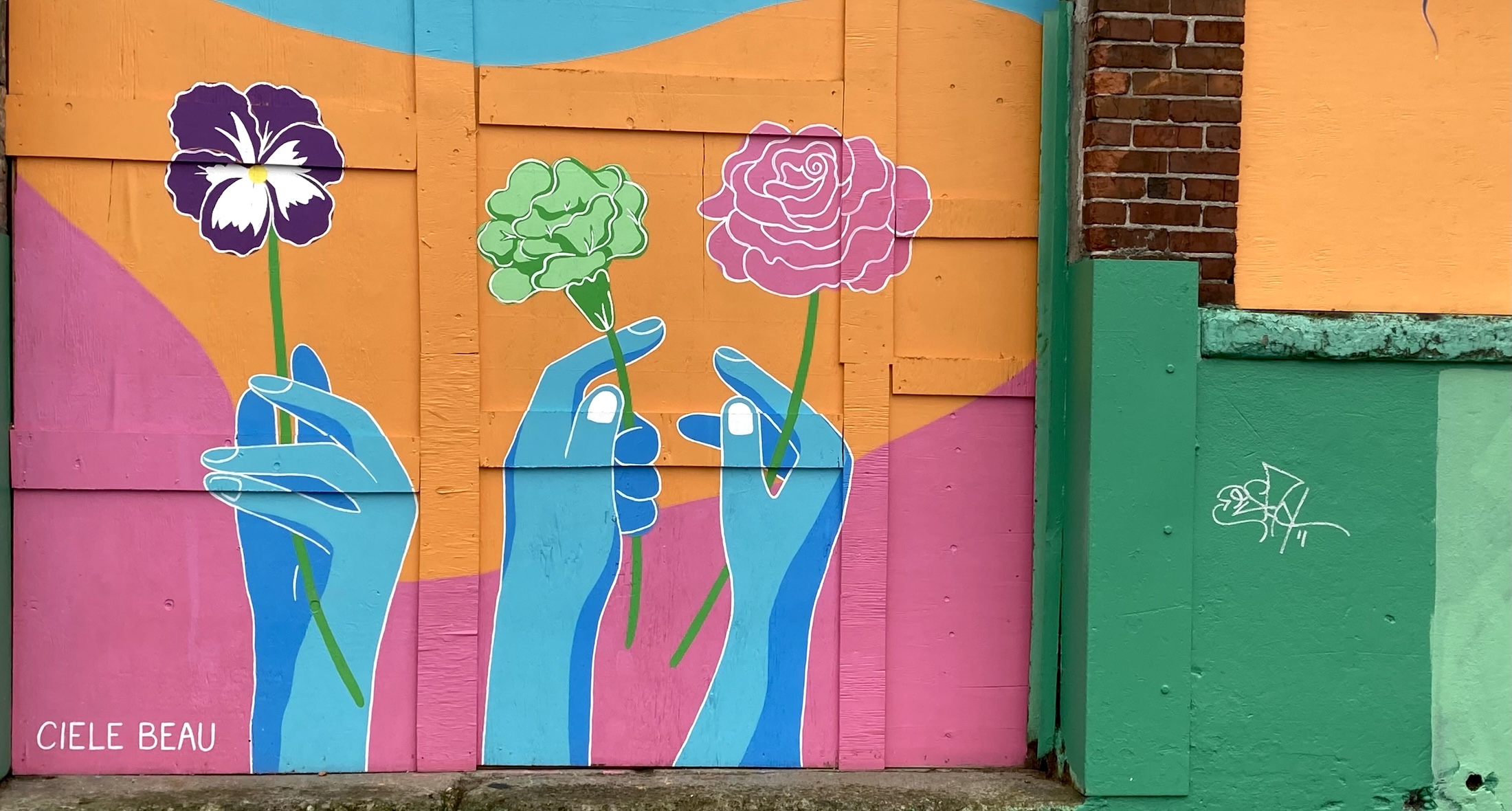 A mural featuring three blue hands holding flowers against an orange, blue and pink background stands beside a brown brick wall painted with bright green trim.