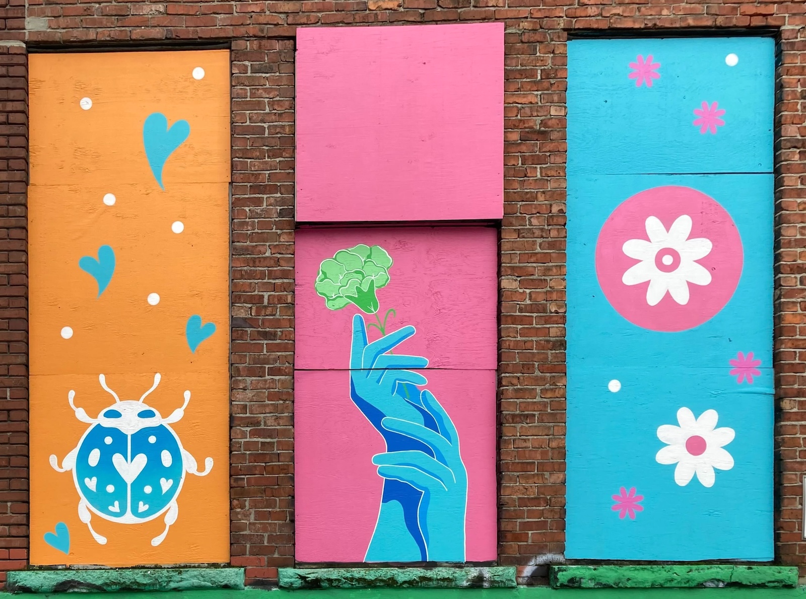 A three-panel mural against a brown brick wall with green ledges features, from left to right, an orange vertical panel with a blue insect; a pair of blue hands holding a green blossom against a pink background; and pink and white flowers against a blue background.
