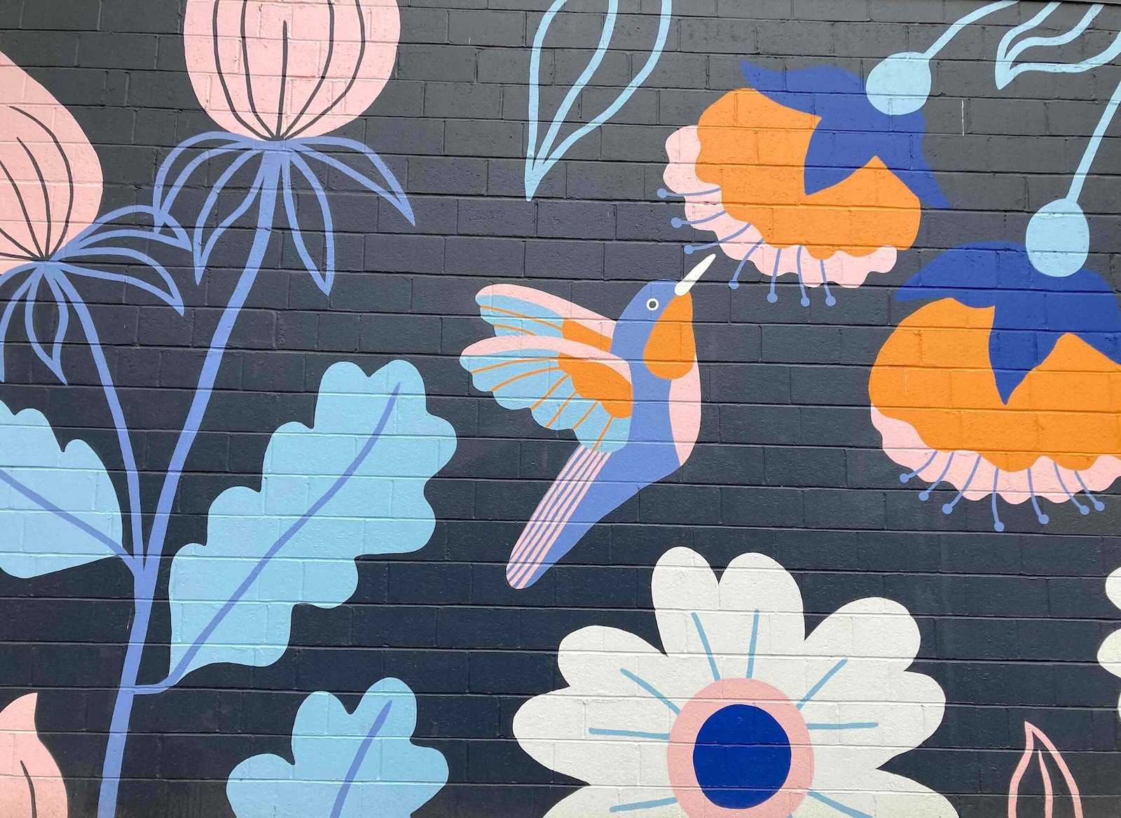 A mural of a hummingbird among flowers in blue and pink tones is painted against a navy background on a brick wall.