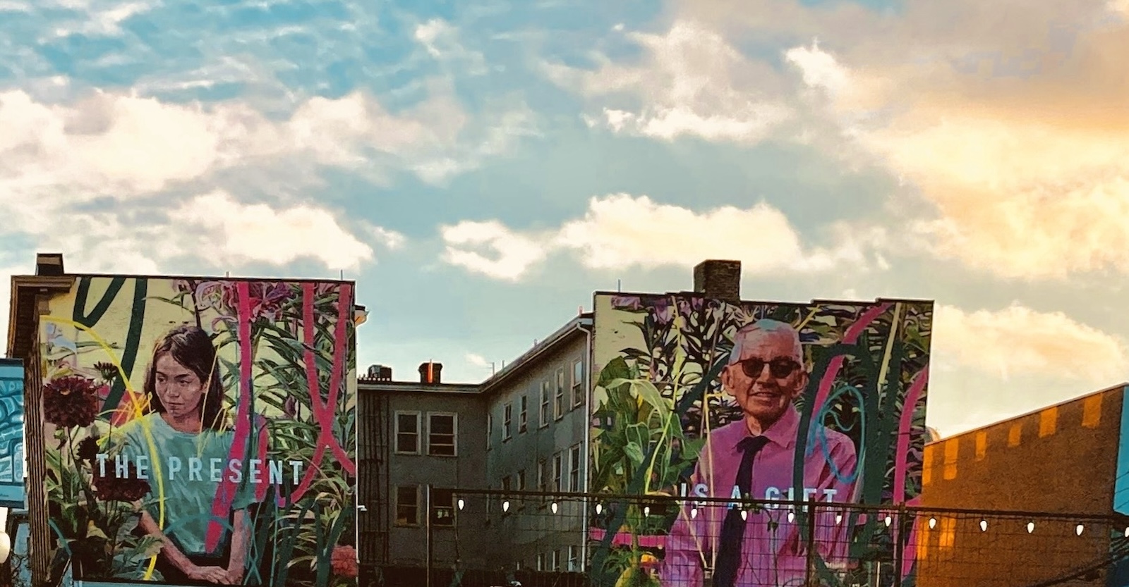 A mural stands across two highrise apartment buildings against a bright sky. On the left is a painting of a girl with brown hair and a green shirt; on the right is a painting of an older man with grey hair, glasses and a pink shirt. The girl’s panel reads 'the present' and the man’s panel reads 'is a gift.'