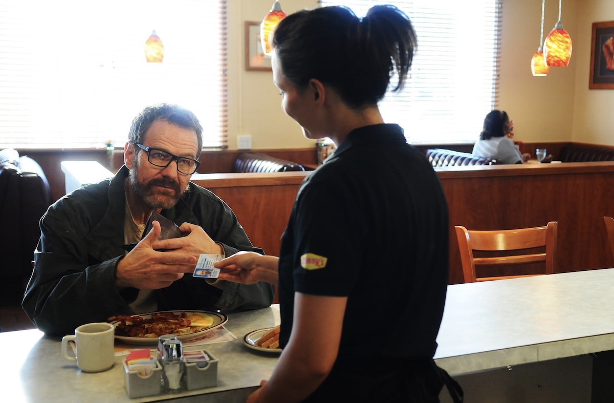 A man with dark hair and glasses sits at the bar in a diner and shows his identification to a waitress, a young woman with dark hair tied up. She is wearing a dark restaurant uniform with a Denny&rsquo;s logo on the sleeve.