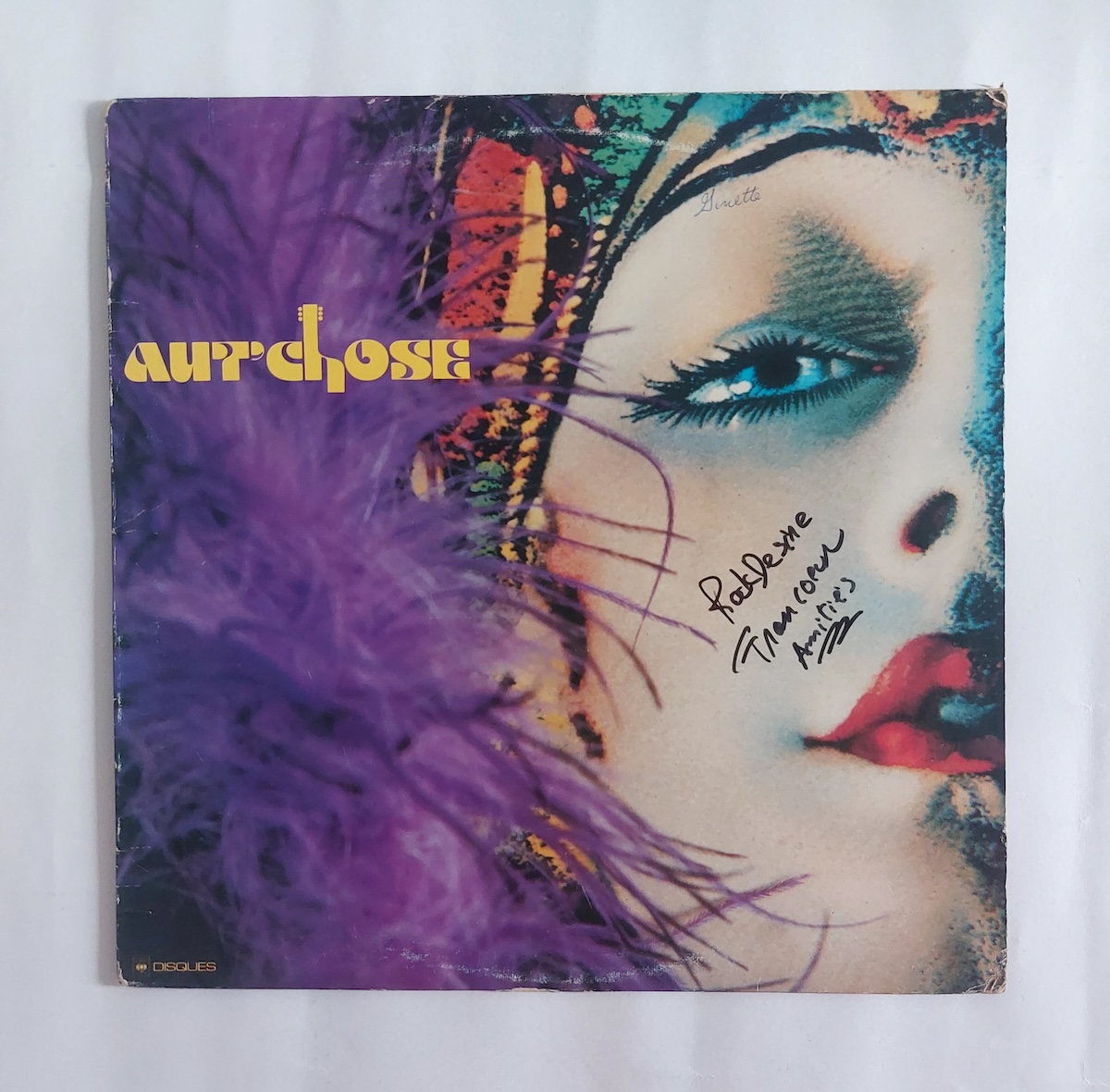 A vinyl album cover features a stylized photo of a woman&rsquo;s face with soft purple feathers and a patterned headpiece on the left side of the frame. She has blue eyes and is wearing red lipstick. Stylized title text in yellow is to the left of the frame.