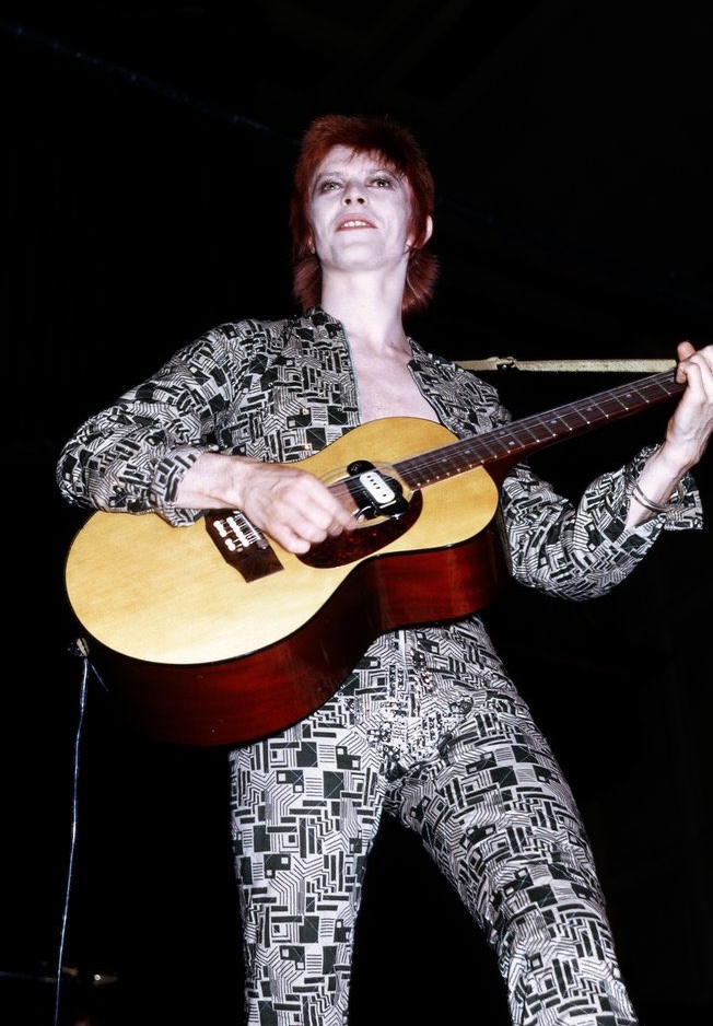 David Bowie performs as Ziggy Stardust and stands holding an acoustic guitar against a black background. He has short red hair and is wearing a black and white patterned jumpsuit.