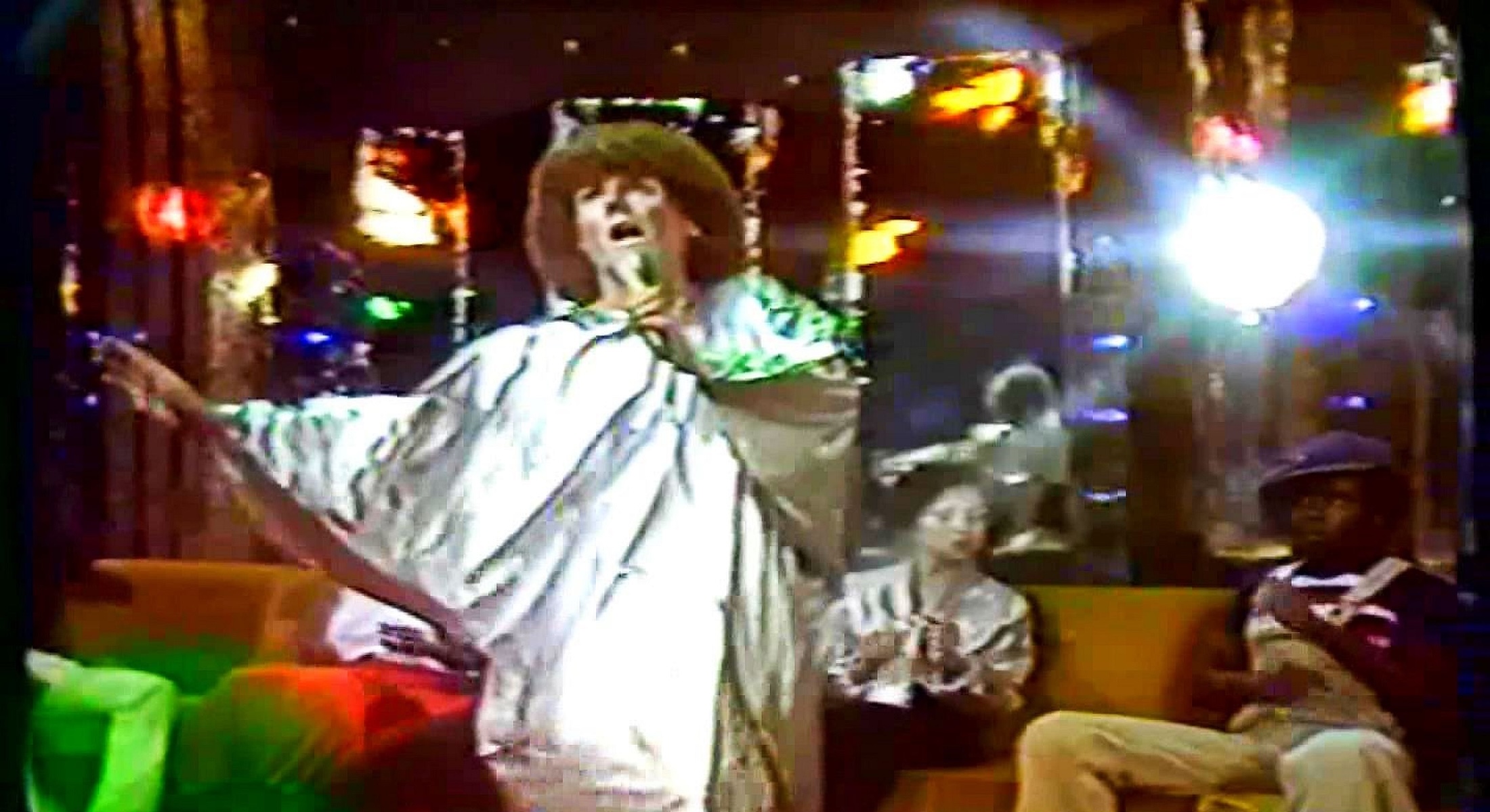 A pixelated colour photograph features a man with brown curly hair and a long white gown singing into a microphone inside a lounge lit with rainbow lights. There are people sitting on the low banquette behind him.